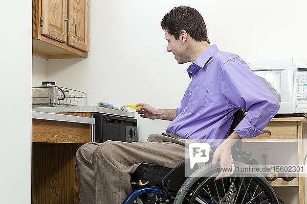 Mann im Rollstuhl mit Querschnittslähmung beim Staubwischen in einer barrierefreien Küche