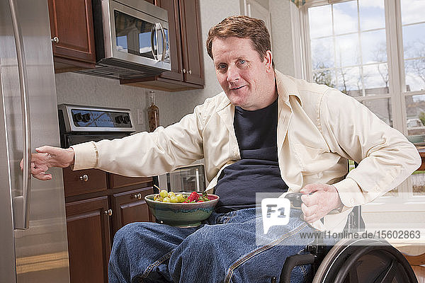 Mann mit Rückenmarksverletzung im Rollstuhl holt Schale mit frischem Obst aus dem Kühlschrank