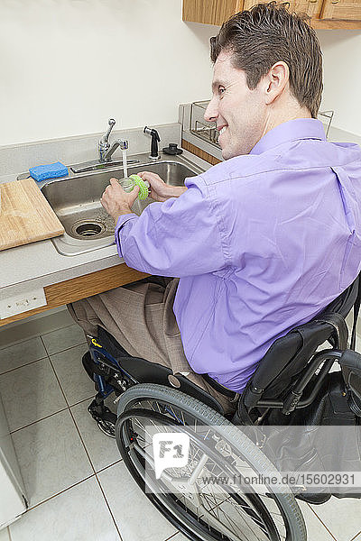 Mann im Rollstuhl mit Rückenmarksverletzung spült Glas in einem begehbaren Spülbecken