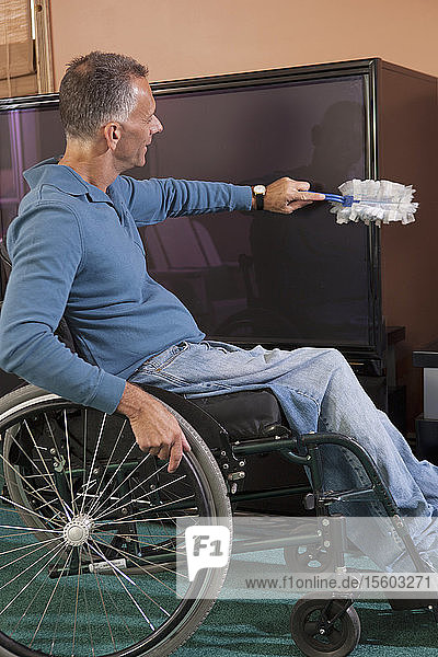 Mann mit Querschnittslähmung im Rollstuhl beim Abstauben von Möbeln