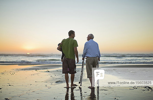 Älterer Mann mit einer Gehhilfe am Strand mit seinem Sohn und seinem Enkelkind