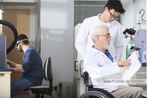 Professor with muscular dystrophy and engineering student reviewing user manual for chemical analyzer in a laboratory