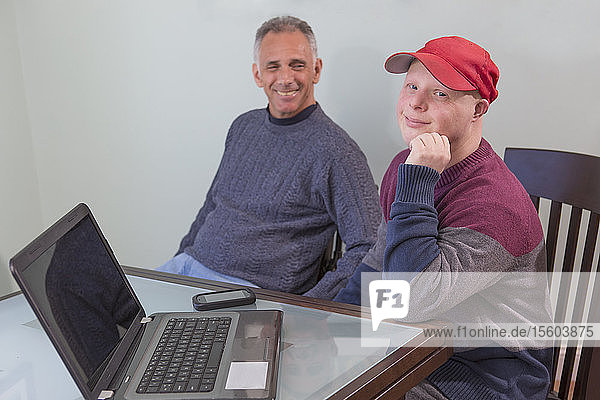 Happy young man with Down Syndrome and his father in wheelchair with Spinal Cord Injury using a laptop at home