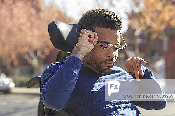 Afroamerikanischer Mann mit Cerebralparese  der draußen auf seinem Elektrorollstuhl eine Brille aufsetzt