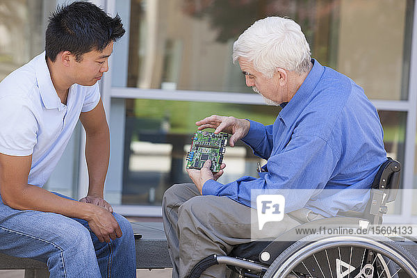 Ingenieur mit Muskeldystrophie und Diabetes in seinem Rollstuhl im Gespräch mit einem Entwicklungsingenieur über Mikrochips auf einer Leiterplatte