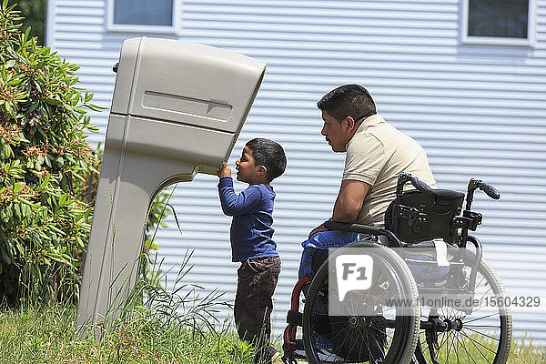 Hispanischer Mann mit Rückenmarksverletzung im Rollstuhl mit seinem Sohn schaut in einen Briefkasten
