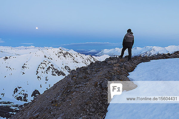 A hiker on a ridge in the Alaska Range looks toward the moon over a series of neighboring ridges after sunset; Alaska  United States of America