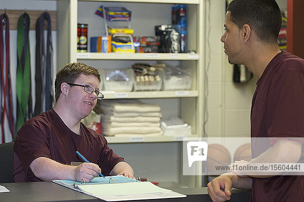 Junger Mann mit Down-Syndrom arbeitet in der Ausgabestelle für Sportgeräte an der Hochschule und spricht mit seinem Vorgesetzten