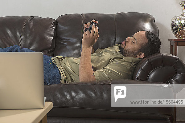 Afroamerikanischer Mann mit Down-Syndrom benutzt zu Hause auf der Couch einen Game-Controller