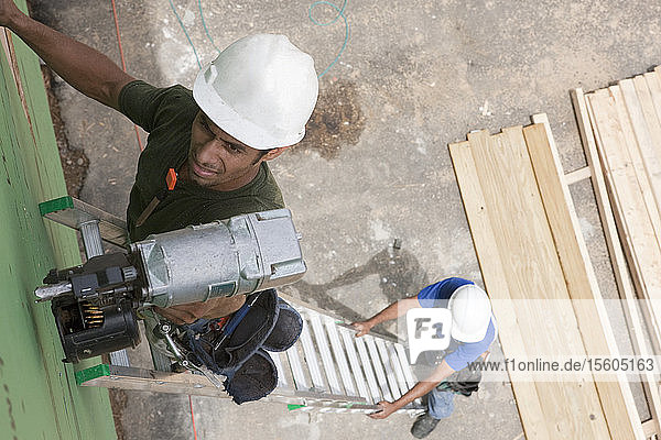 Spanischer Zimmermann  der auf einer Leiter eine Nagelpistole benutzt und von einem anderen Zimmermann gestützt wird