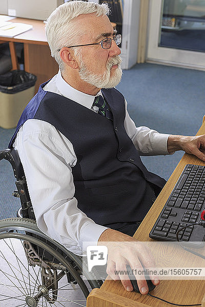 Mann mit Muskeldystrophie im Rollstuhl bei der Arbeit an seinem Computer in einem Büro