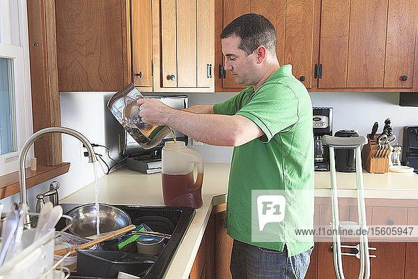Mann nach Operation des vorderen Kreuzbandes (ACL) mit Krücken beim Teekochen und Abwaschen in der Küche