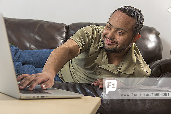 Glücklicher afroamerikanischer Mann mit Down-Syndrom  der zu Hause vom Sofa aus einen Laptop benutzt