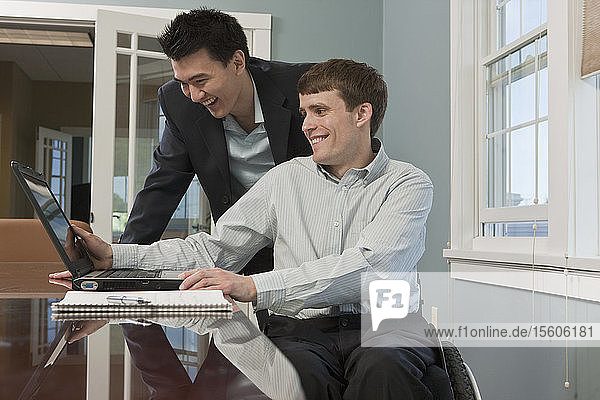 Ein querschnittsgelähmter Geschäftsmann benutzt mit seinem Kollegen in einem Büro einen Laptop