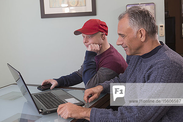 Junger Mann mit Down-Syndrom und sein Vater mit Rückenmarksverletzung benutzen zu Hause einen Laptop