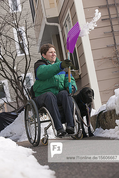 Frau mit Multipler Sklerose schaufelt Schnee im Rollstuhl mit einem Diensthund