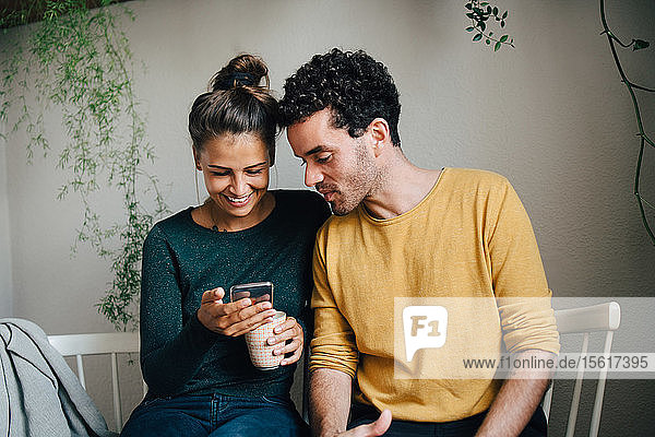 Smiling girlfriend showing smart phone to boyfriend while having coffee in living room