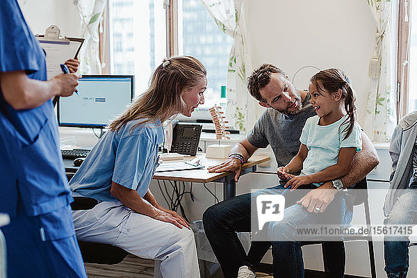 Female pediatrician talking to smiling girl sitting with father in medical examination room