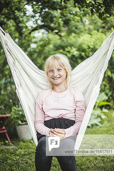 Porträt eines lächelnden Mädchens  das am Wochenende auf einer weißen Schaukel im Hinterhof sitzt