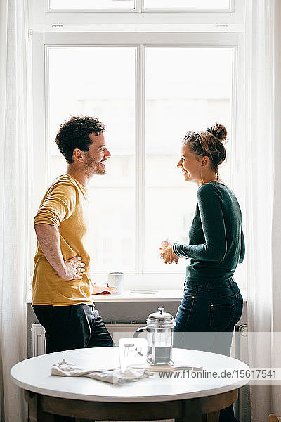 Seitenansicht eines glücklichen Paares  das sich unterhält  während es zu Hause am Fenster steht