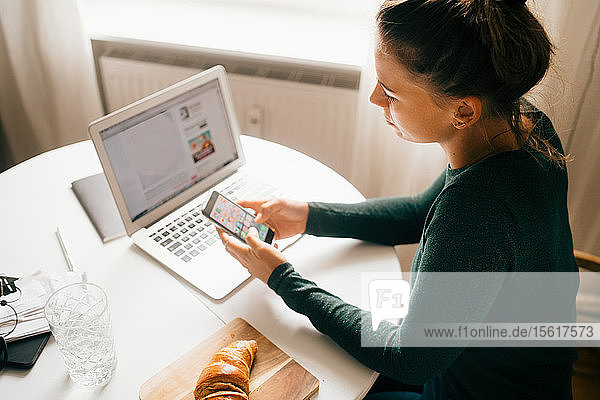 Frau benutzt Smartphone  während sie zu Hause mit ihrem Laptop auf einem Lebensmittelrezept surft