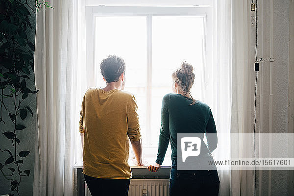 Rückansicht eines zu Hause stehenden Paares  das durch ein Fenster schaut