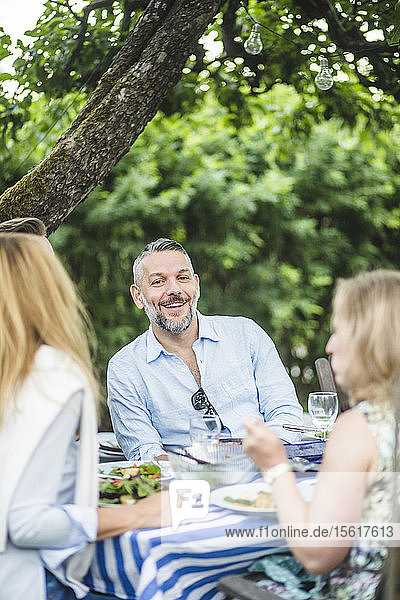 Glücklicher Mann genießt mit Freunden am Esstisch bei einer Gartenparty