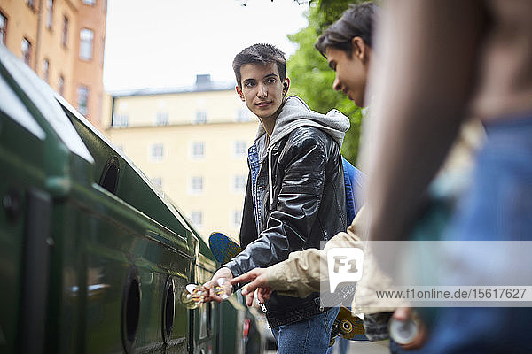 Teenager  der einen Freund ansieht  während er Abfälle in den Mülleimer einer Recycling-Station wirft