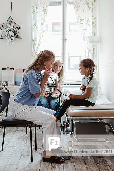 Kinderarzt hört den Herzschlag des Mädchens durch ein Stethoskop ab  während die Mutter mit dem Jungen im Hintergrund in der Klinik sitzt