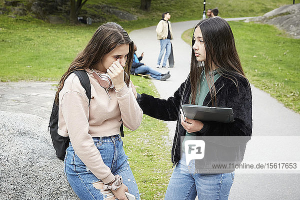 Teenage girl looking at female friend crying while sitting on rock at park