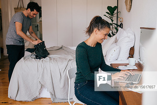 Smiling woman using laptop while boyfriend looking in purse at home