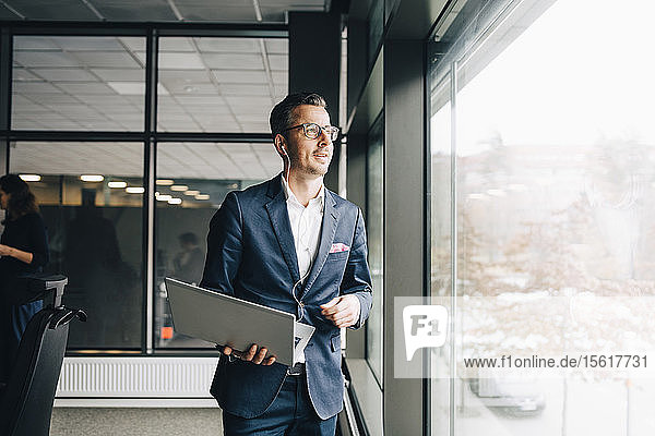 Lächelnder Unternehmer hält Laptop in der Hand  während er im Büro durchs Fenster schaut