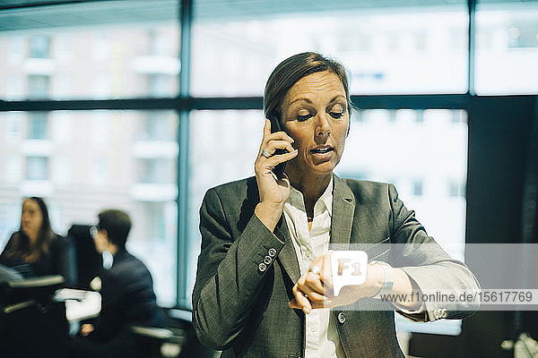 Vielbeschäftigte Geschäftsfrau telefoniert mit dem Handy  während sie im Büro die Zeit auf die Uhr schaut