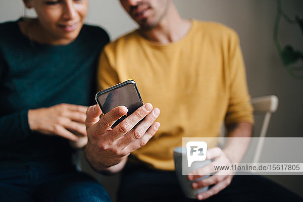 Midsection of boyfriend showing smart phone to girlfriend while having coffee at home