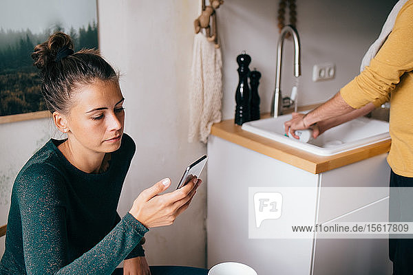 Mid adult woman using mobile phone while boyfriend washing utensils at home
