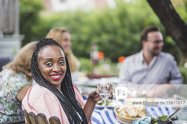 Porträt einer lächelnden Frau  die Wein trinkt  während sie mit Freunden auf einer Gartenparty sitzt