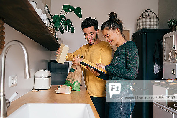 Lächelnder Mann nimmt Essen aus Tüte  während seine Freundin ein digitales Tablett an der Küchentheke zeigt