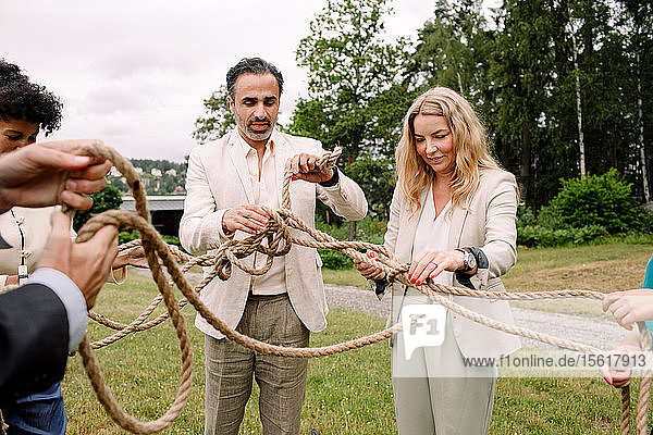 Male and female business professionals holding tangled rope while standing in lawn