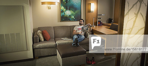 Ein smarter junger Mann sitzt in einem Hotelzimmer auf einem Sofa  hat die Beine auf einen Hocker gelegt und liest eine Zeitung.