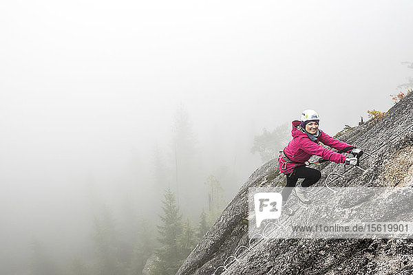 Eine Frau lächelt aufgeregt  als sie die Metallsprossen eines Klettersteigs in Squamish hinaufklettert.