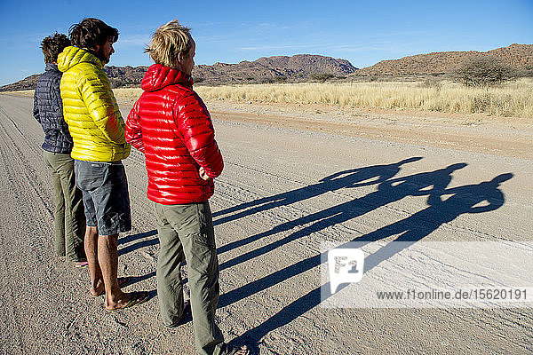 Drei Männer in Jacken werfen lange Schatten auf eine unbefestigte Straße  Erongo-Region  Namibia