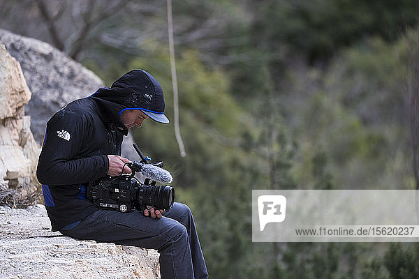 Männlicherï¾ Videofilmerï¾ sitzt auf einer Felsformation  richtet seine Kamera und filmt etwas in der Ferne  ï¾ Siurana  Katalonien  Spanien