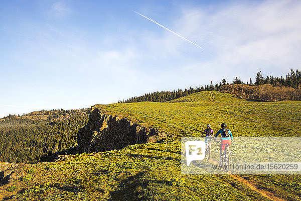 Two young women ride mountain bikes on a single-track trail through green grass in early morning sunlight.