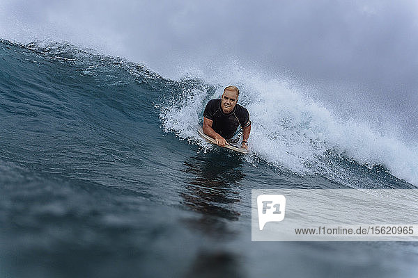 Männlicher Surfer schaut in die Kamera  während er auf dem Surfbrett liegt und eine Welle reitet  Male  Malediven