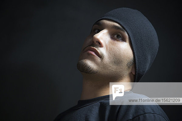 Low angle portrait of a young Hispanic man with a black cap pulled down on his head.
