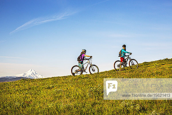 Two young women push mountain bikes up a single-track trail through an open meadow.