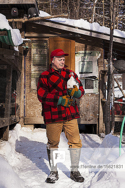A portrait of a farmer looking affectionately at a guinea hen in front of a chicken coop on a winter's day.