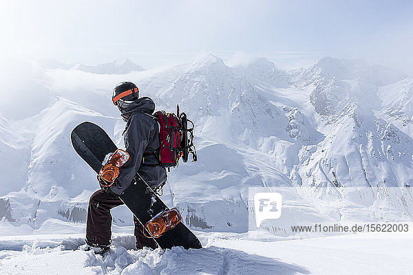 Ein Backcountry-Snowboarder mit seinem Board steht auf einem Bergrücken und überblickt die entfernten Berge