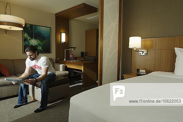 Ein smarter junger Mann sitzt auf einem Hocker in einem Hotelzimmer und liest eine Zeitung.