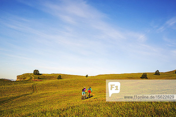 Zwei junge Frauen fahren mit dem Mountainbike auf einem einspurigen Weg durch grünes Gras in der frühen Morgensonne.